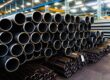 Large and small steel pipes are stacked in organized bundles inside a manufacturing facility. The ends face the viewer.