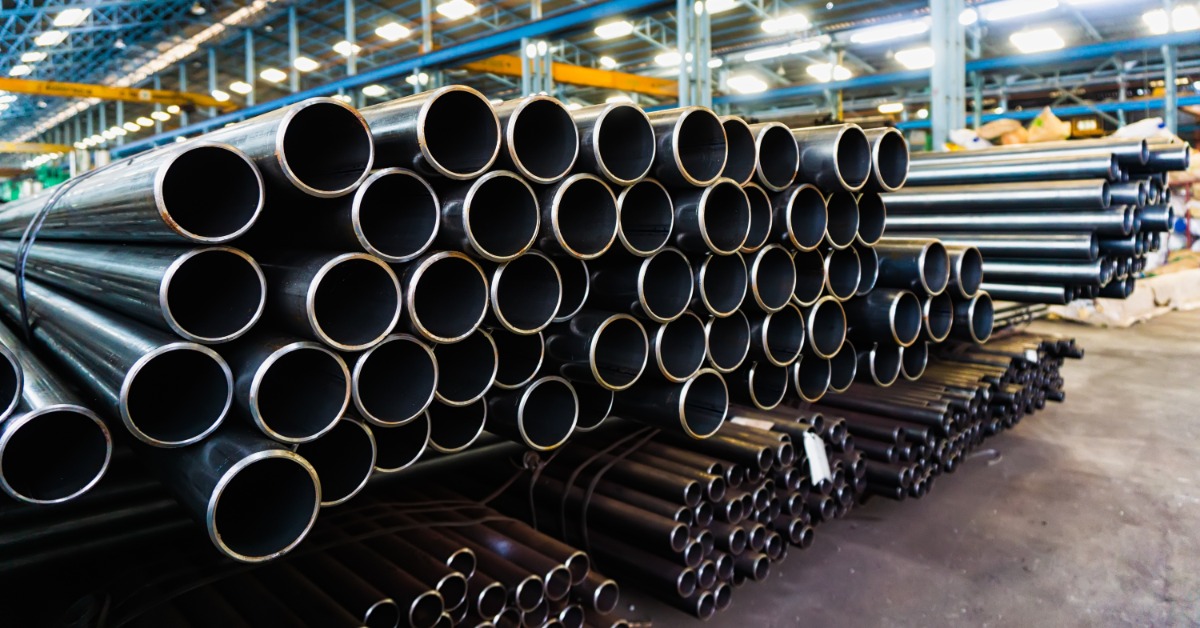 Large and small steel pipes are stacked in organized bundles inside a manufacturing facility. The ends face the viewer.