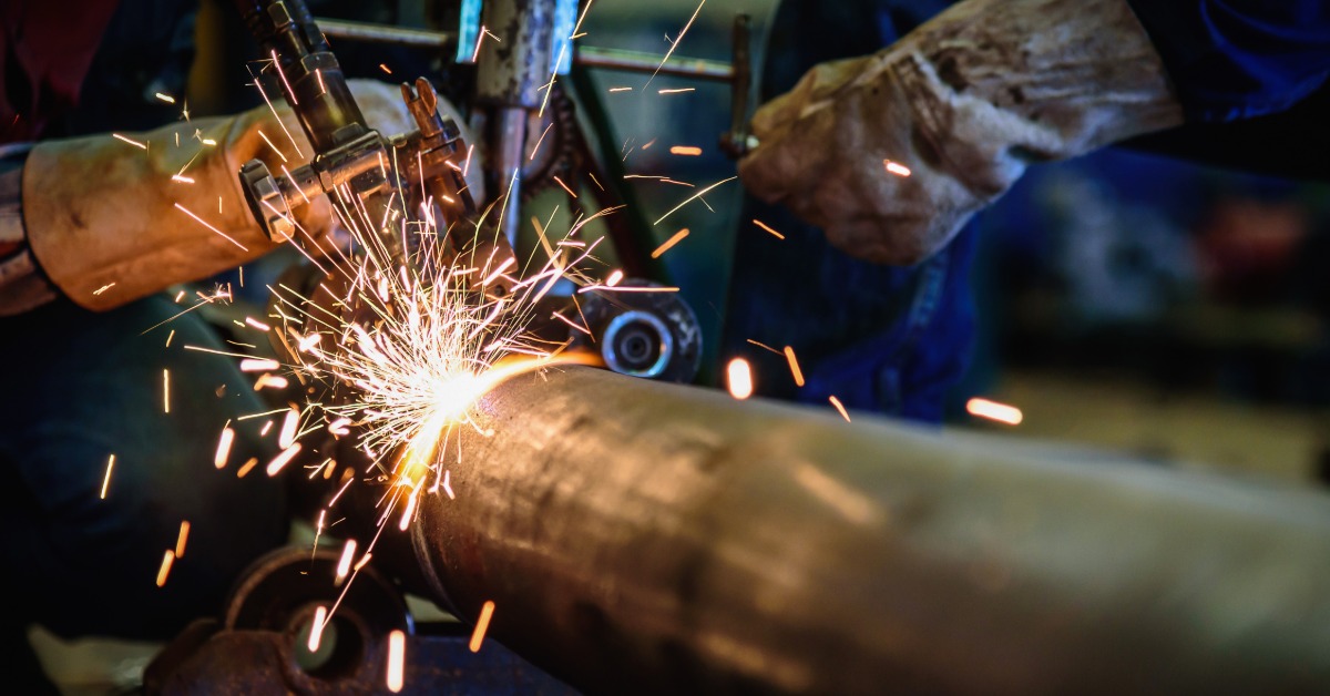 A worker welds a large steel pipe while wearing protective gloves. Bright orange sparks scatter from the welding point.