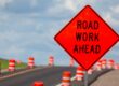 An orange diamond-shaped “Road Work Ahead” sign is beside a roadway. Orange-and-white traffic barrels line the road.