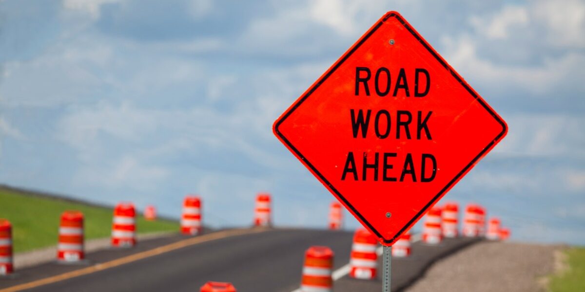 An orange diamond-shaped “Road Work Ahead” sign is beside a roadway. Orange-and-white traffic barrels line the road.