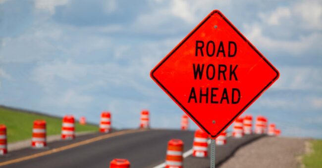 An orange diamond-shaped “Road Work Ahead” sign is beside a roadway. Orange-and-white traffic barrels line the road.