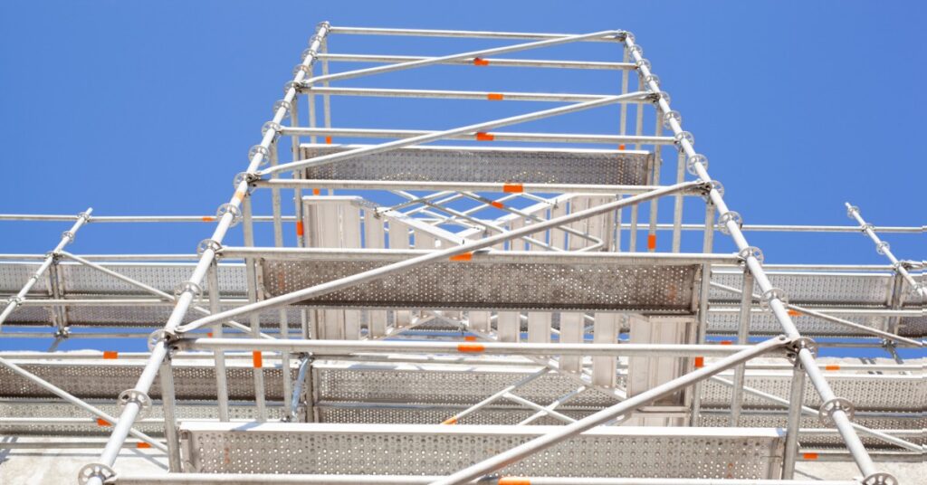 A tall metal scaffolding structure rises against a blue sky. Silver tubes, braces, and platforms form the framework.