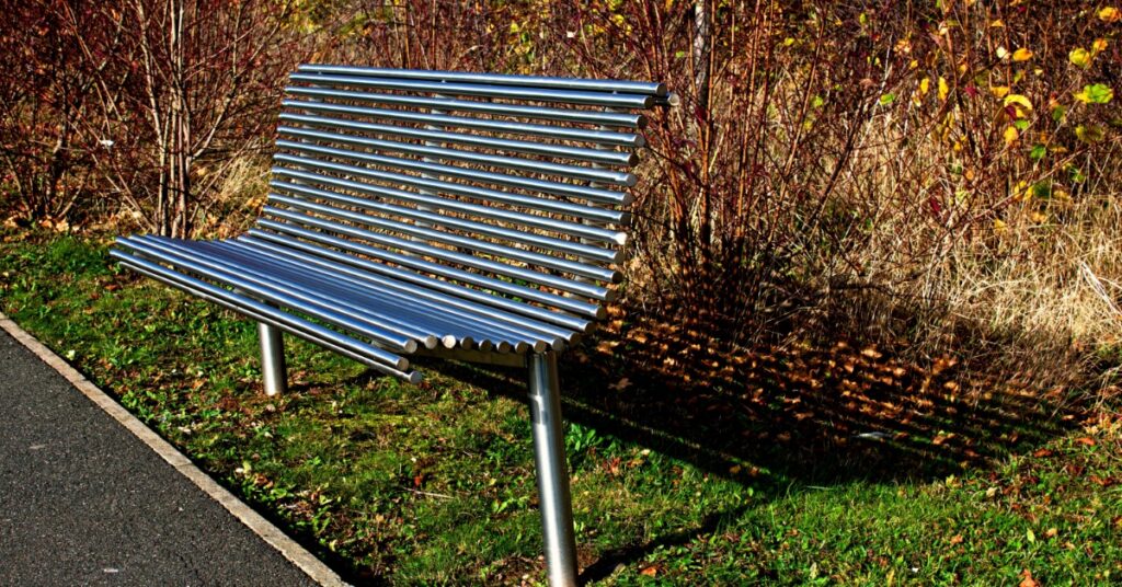 A metal bench made from parallel silver tubes sits beside a paved path. Autumn-colored foliage surrounds the bench.